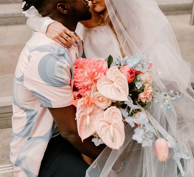 Bride with a long veil and holding a pink and blue wedding bouquet sitting on her grooms lap in a patterned shirt 