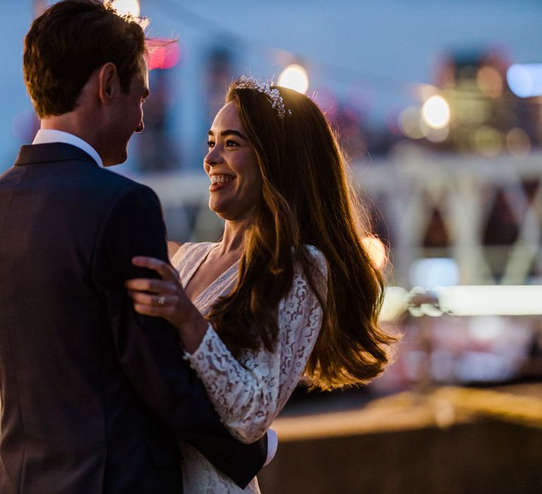 Bride in lace sheer sleeved wedding dress and pearl cluster headband with long brown hair smiles at groom