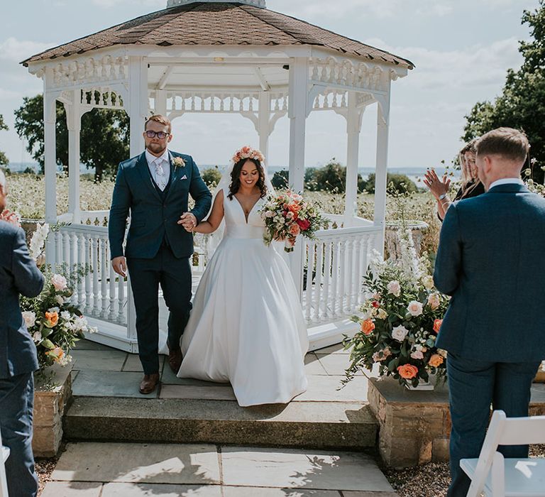 Bride and groom exit their outdoor wedding ceremony together with groom in three piece blue suit with white floral tie and bride in Stella York dress