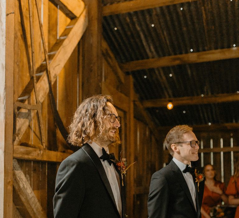 Groom and groomsmen get their first look at the bride in black tie, black glasses and orange dried flower buttonhole