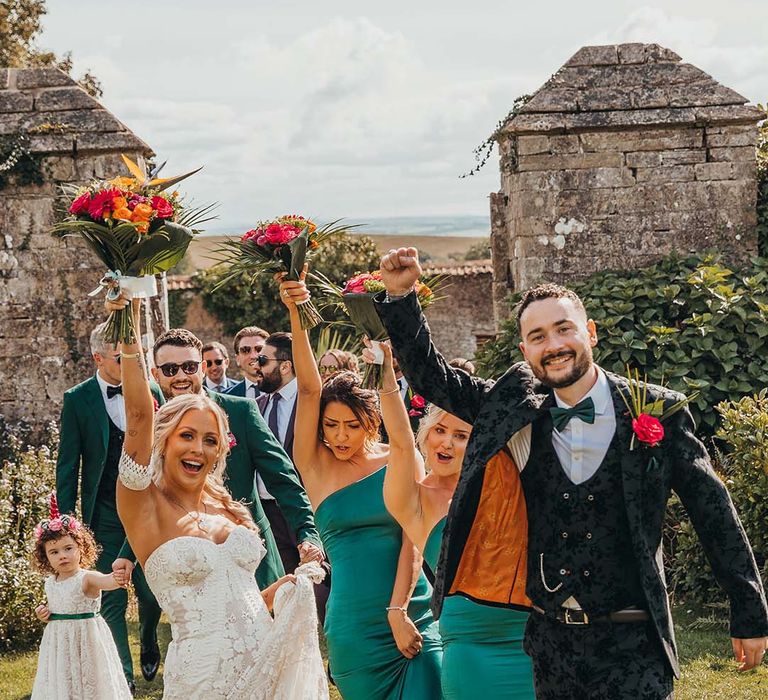 Bride in boho lace wedding dress and blue wedding shoes lifts bouquet in celebration as groom, bridesmaids and other guests fist pump