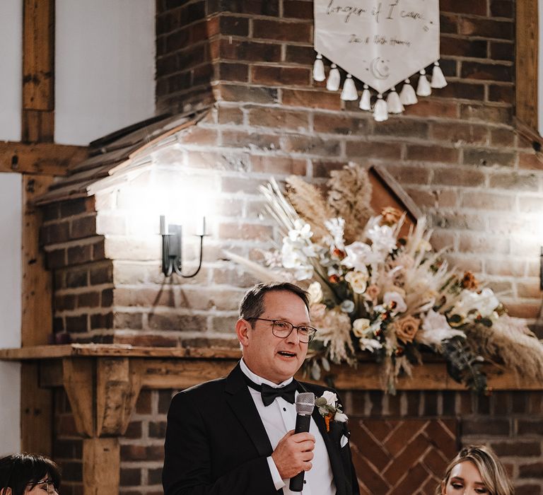 Father of the bride in black tie gives a speech with boho dried flower large display decor on fireplace 