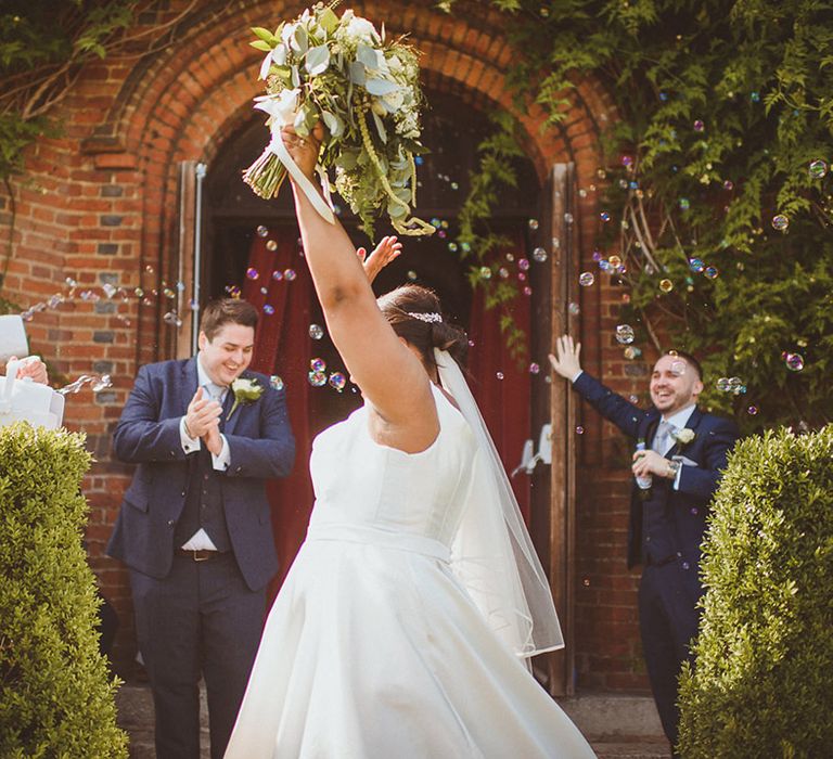 Bride raises her arms and bouquet as groomsmen use bubble machine to blow out bubbles 