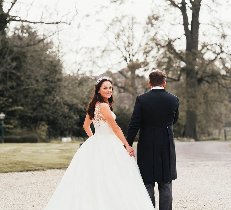 Bride in princess Pronovias wedding dress looks back at the camera walking holding hands with groom 