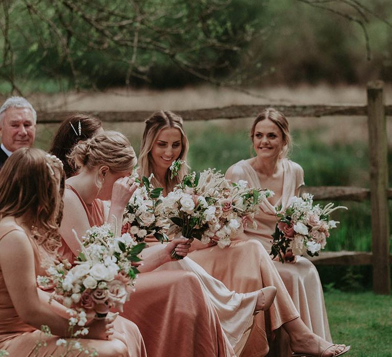 Bridesmaids smile and wipe their eyes during the wedding ceremony all holding matching bouquets with pink and white roses and dried grass