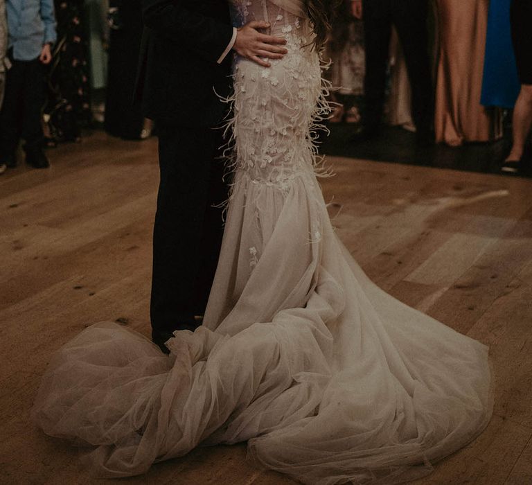 Bride in floral wedding dress with feathers and tulle skirt with groom in blue suit as they share their first dance with wedding guest watching and filming 