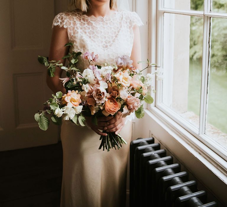 Blonde haired bride stands beside window wearing Kate Beaumont gold silk wedding gown complete with pastel coloured floral bouquet 