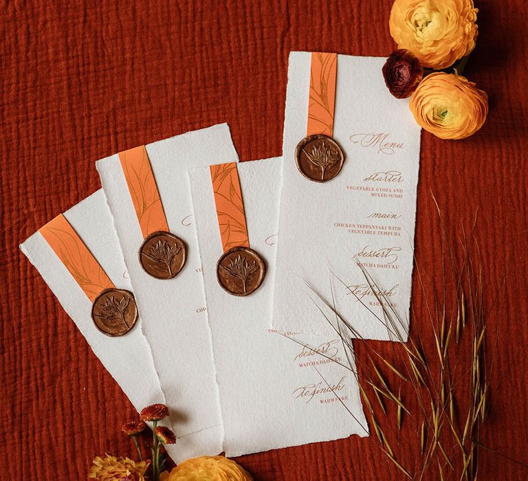 Menu cards written in gold calligraphy with orange design held on by a copper wax seal 