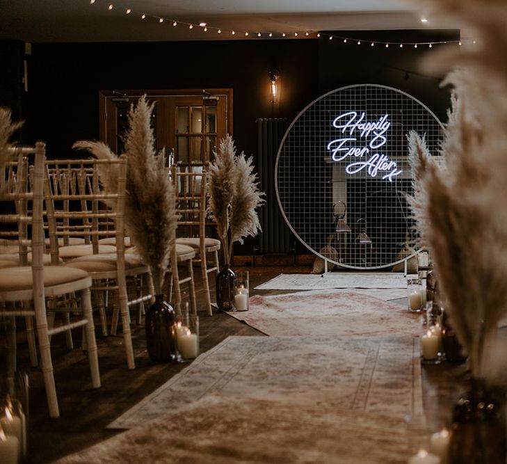 Pampas grass lines aisle alongside white chairs complete with neon sign and Moroccan styled rugs 