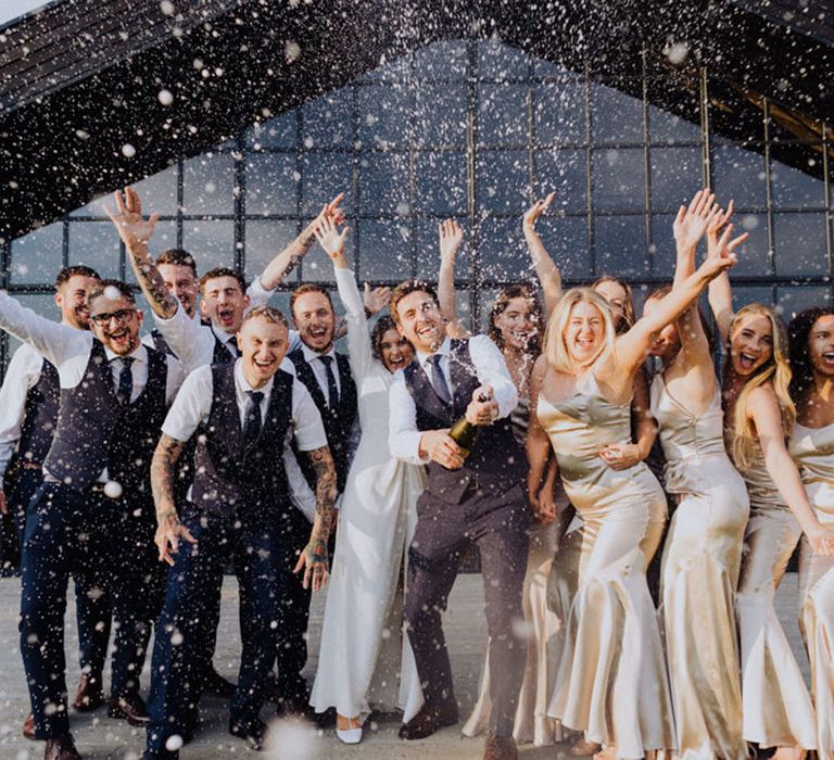 Bride and groom pose with the groomsmen and bridesmaids in gold dresses pop champagne bottles 