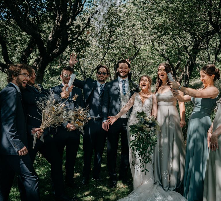 Bride wearing lace wedding dress stands with her groom and bridesmaids who wear sage green dresses in differing styles as they let off confetti cannons with the groomsmen