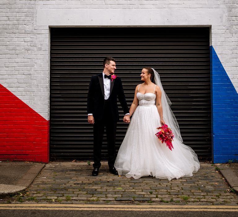 Bride wearing fitted sparkly wedding dress & groom in velvet jacket stand in front colourful wall outdoors in Brixton 