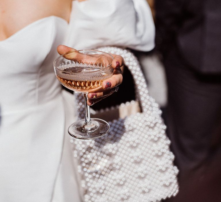 Bride with sparkly pink heart wedding nails holding a coupe glass with champagne and a pearl handbag 