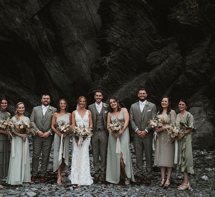 Bride & groom stand with their bridesmaids who wearing different styled green bridesmaid dresses and grooms in three piece suits at Tunnels Beach 