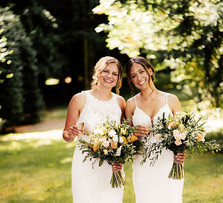 Brides stand together with their champagne glasses with a white and orange flower bouquet in fitted lace wedding dresses