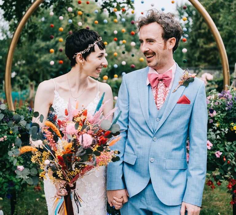 Bride holds colourful dried floral bouquet and wears bridal crown alongside her groom in dusty blue suit with pink ruffled shirt and bow tie