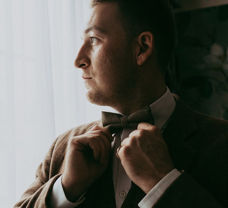 Groom looks out of the window as he does his bow tie for the classic style wedding 