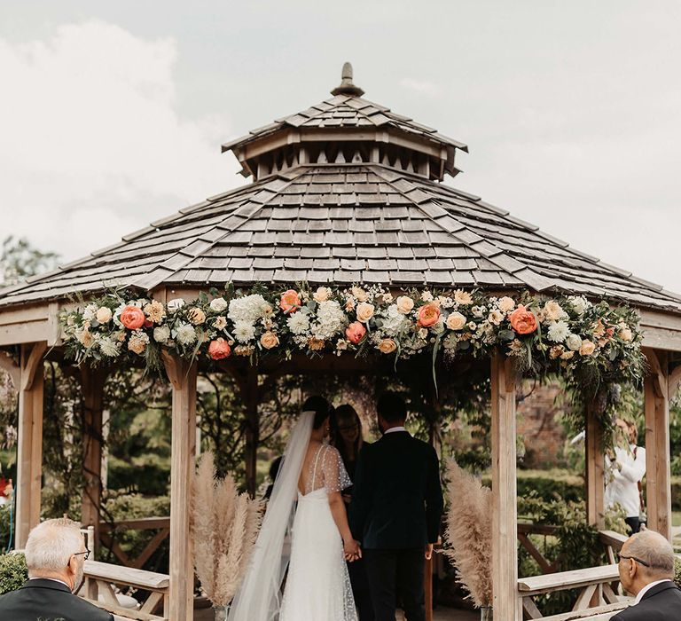 Bride and groom stand in the gazebo with their hands intertwined as they participate in their outdoor wedding ceremony 