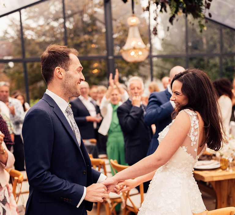 Bride and groom stand holding hands as the wedding guests clap and applaud 