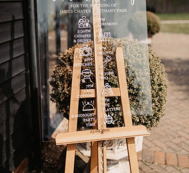 Clear acrylic order of the day wedding sign on a wooden easel with foliage decoration 