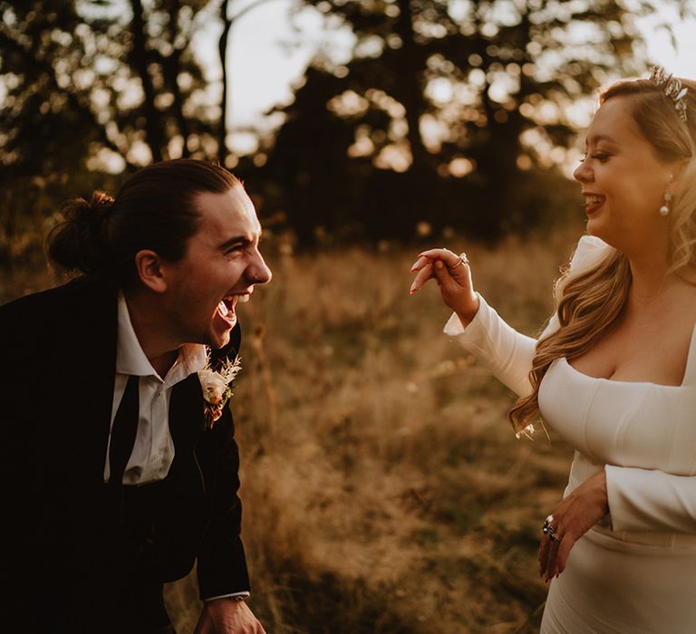 Groom in black suit with floral boutonniere laughing with bride in square neck corset look satin wedding dress