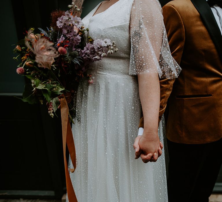 The couple holding hands whilst showing off the bridal bouquet which is tied with a burnt orange ribbon and features Thistles, dried grass and lilac Delphiniums