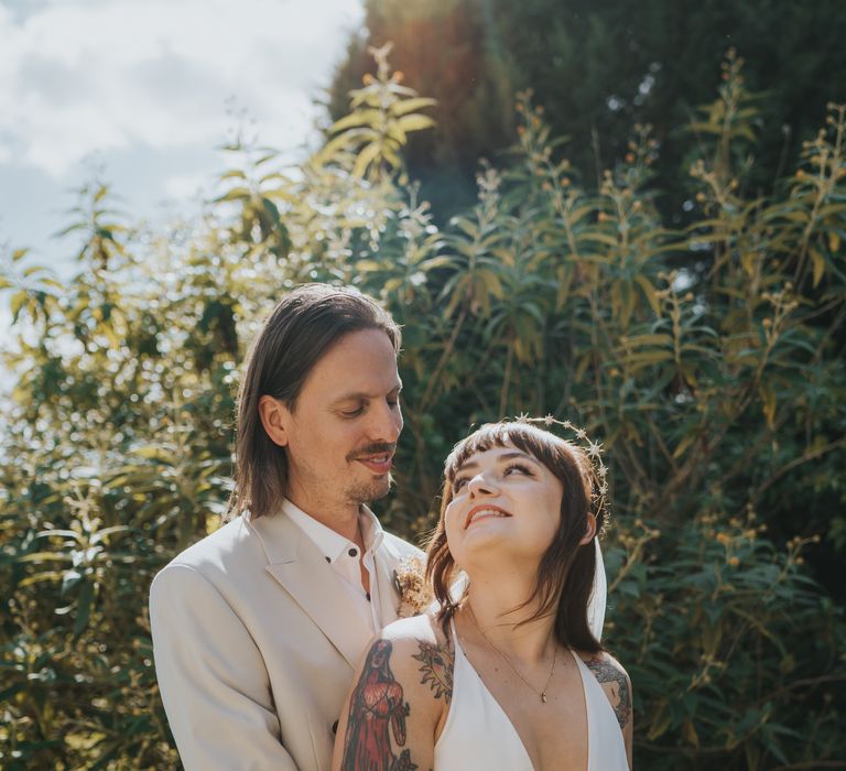 Groom in neutral coloured double-breasted ASOS suit and black high-top converse with dried flower and foliage boutonniere and bride in silk-look v-neck sleeveless ASOS wedding dress and gold halo headband with sparkly stars posing together after wedding at The Reid Rooms