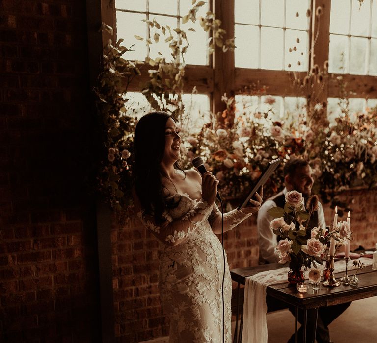 Bride in off the shoulder Abigail Westrup bridal gown with long, sheer sleeve and lace detailing at The Giraffe Shed wedding venue giving her bride's speech to wedding guests and husband