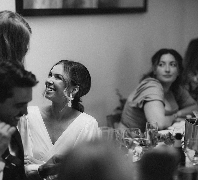 Bride in statement earrings smiles towards wedding guest during intimate reception