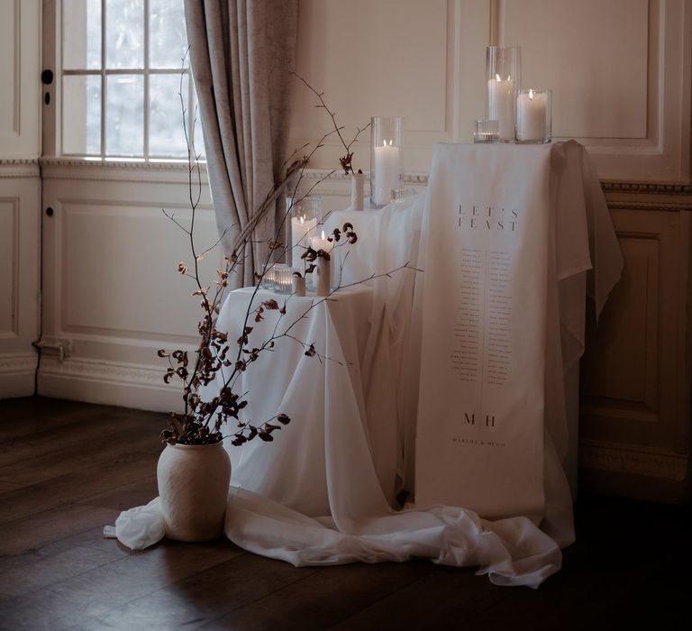 Minimalist wedding decor at Eaves Hall Clitheroe - pillar candles in large glass candle jars on tables with different heights, long satin look wedding drapes, minimalistic wedding sign and dark dried flowers in ceramic vase 