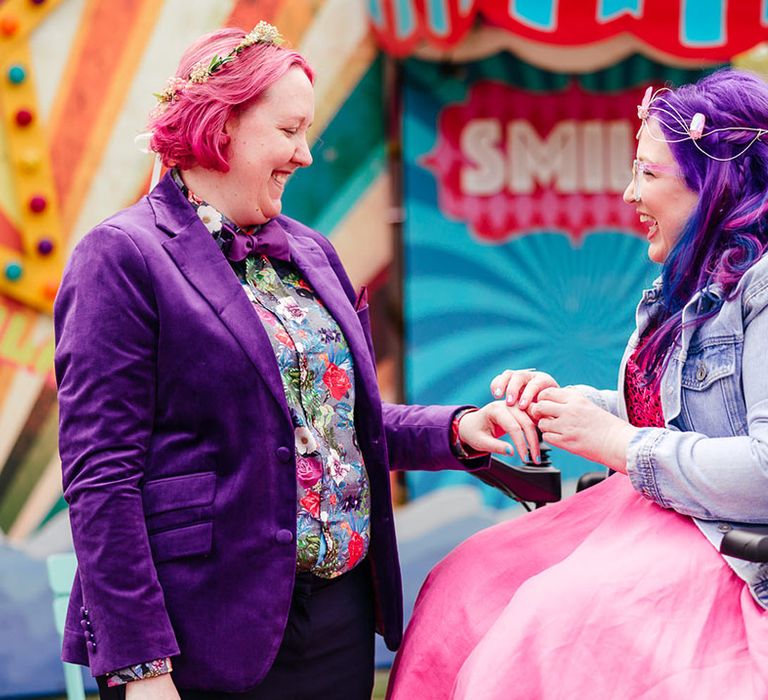 Bride in purple velvet blazer and black suit trousers with purple bowtie, pink patterned pocket square and colourful patterned shirt and bridal flower crown with bride in wheelchair wearing personalised denim jacket, pink wedding dress and gemstone bridal crown 