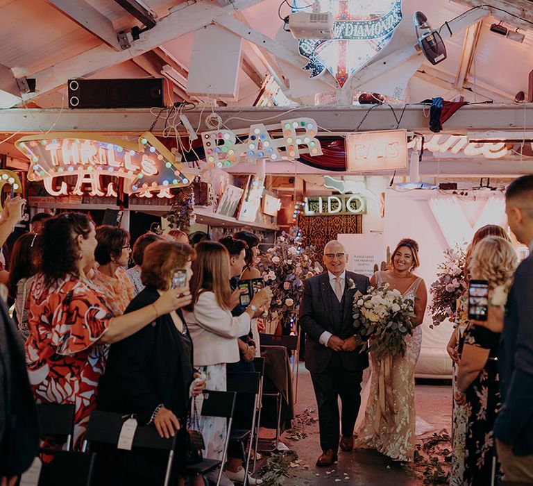 Bride in sleeveless plunge lace wedding dress  and cathedral length embroidered veil with large white rose and foliage bouquet walking down the aisle with father of the bride wearing dark suit with pink tie 