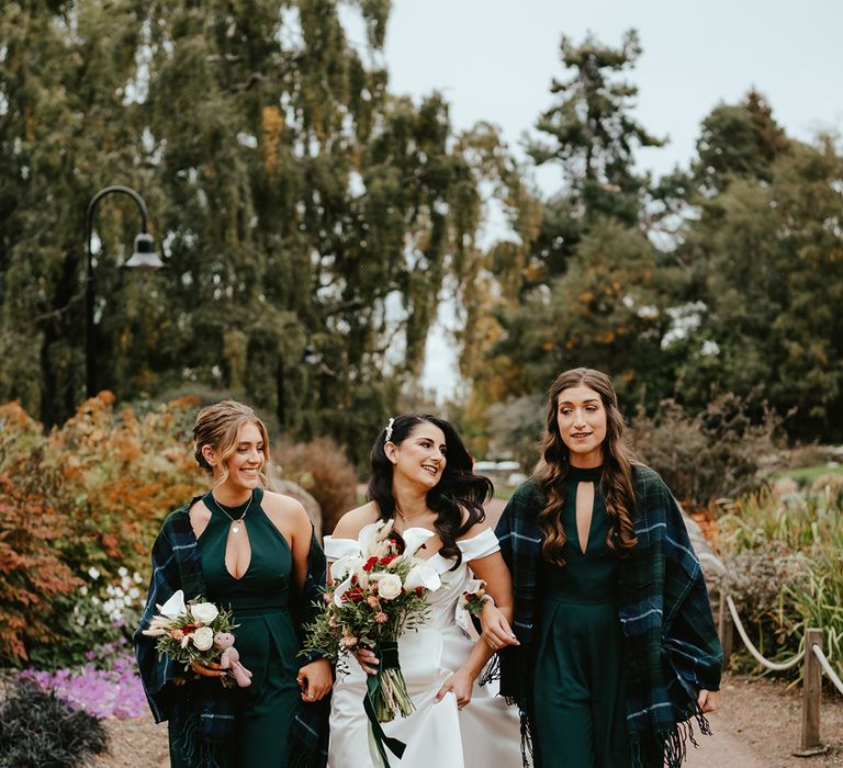 Bride stands with her bridesmaids who wear green bridesmaid jumpsuits with high-necks and tartan shawl coverups 