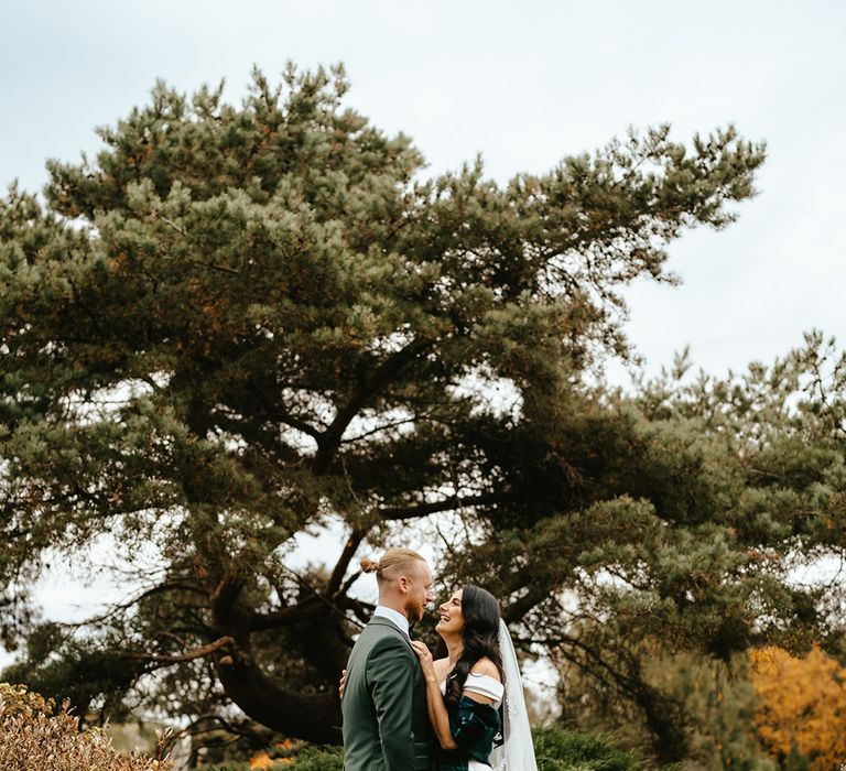 Bride wears tartan shawl coverup and leans into her groom in green suit during outdoor couples portraits 