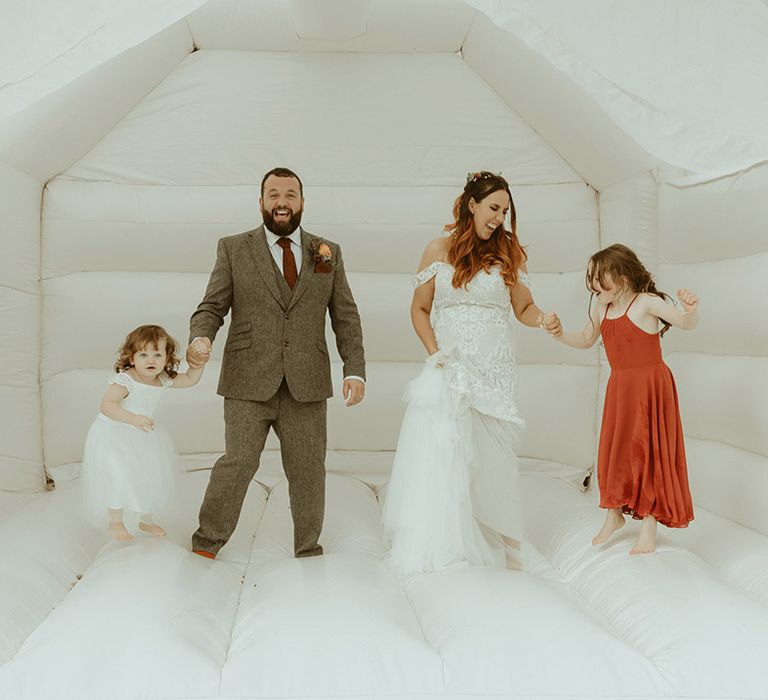 Groom in grey tweed suit with burgundy tie holding hands with daughter in white tulle dress with the bride in boho fitted lace wedding dress holding hands with daughter in burgundy dress for bouncy castle wedding 