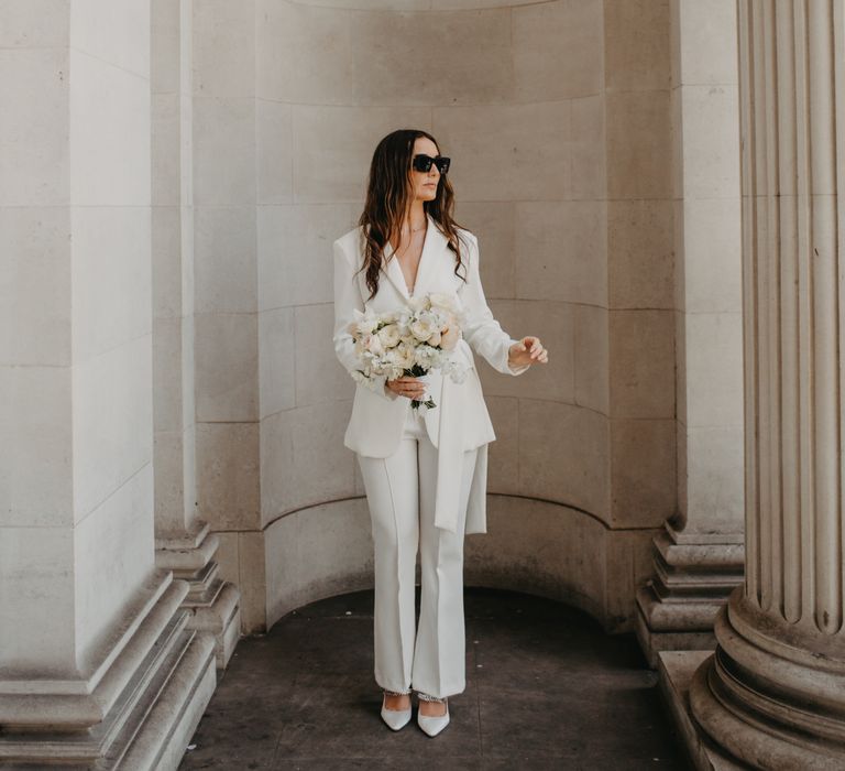 Bride wears fitted bridal suit with tie to waist and tapered trousers with flared bottoms 