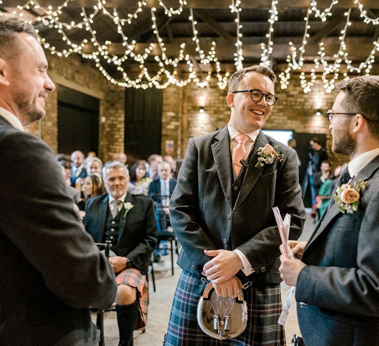 Groom in grey jacket and pale pink tie with tartan kilt during Christian wedding ceremony 