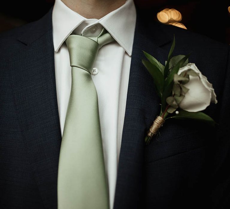 Close up of the groom in a navy suit with a sage green tie and white rose buttonhole 