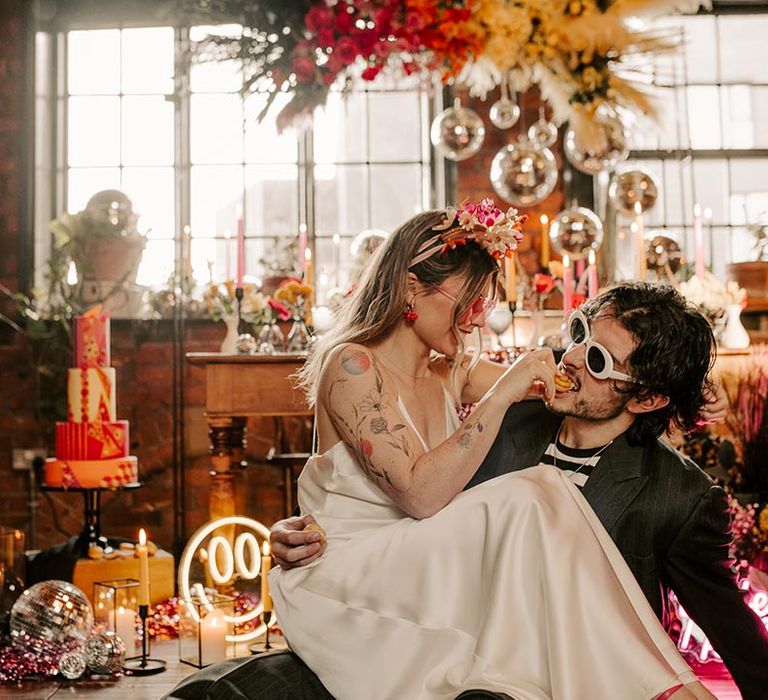 Bride in satin v-neck wedding dress feeding groom in pinstripe suit and black and white shirt a cookie sitting in front of colourful wedding tablescape with pink metallic streamers, hanging disco ball decorations, yellow smiley face neon sign, viva magenta, fuchsia and hot pink tapered candles and suspended pink and yellow rose flowers with foliage 