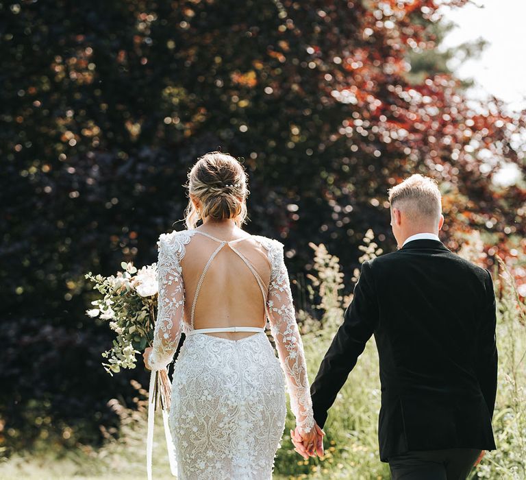 Bride in long sleeve shoulder pad flower lace wedding dress with open back design with the groom hand in hand in black tie 