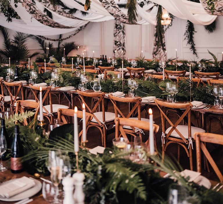 Leaf patterned fabric with foliage and chandeliers hanging from the ceiling with long banquet tables covered in leafy arrangements 