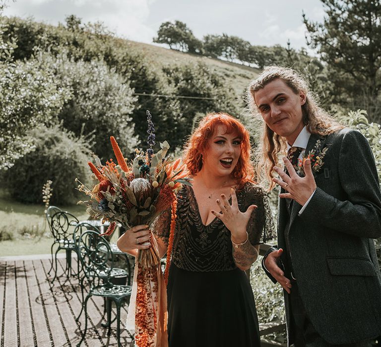 Bride in detailed, embellished v-neck black dress for wedding in Devon, standing with groom in dark grey three piece suit and burgundy flower patterned tie