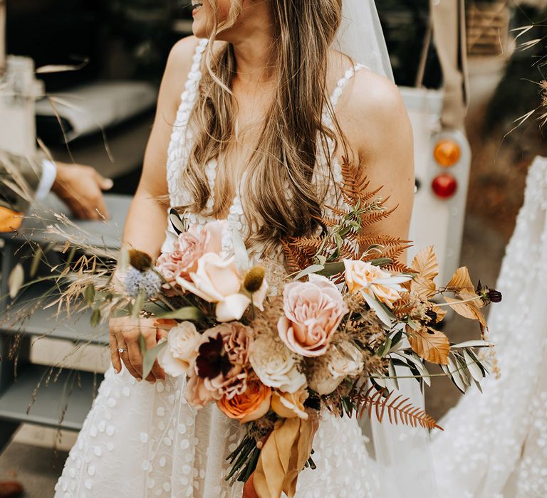 The bride with a braided half up half down wedding hairstyle holding an autumnal pink rose and orange autumnal wedding bouquet 