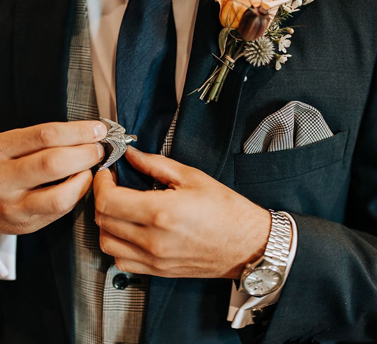 A silver bird tie clip worn by the groom with checkered waistcoat and pocket square and blue suit 