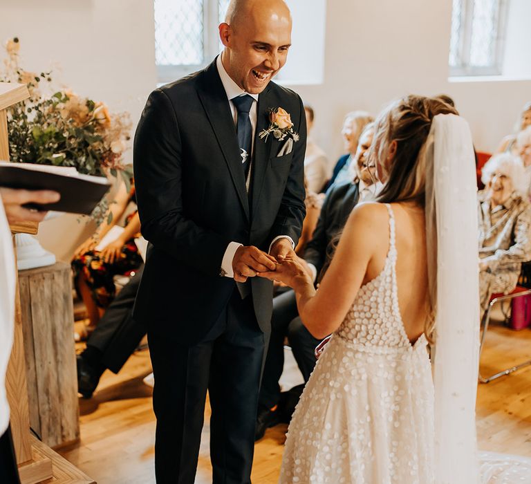 Groom in suit with blue tie and silver bird tie clip puts the wedding ring on the bride's finger with her hair in a braided half up half down style  