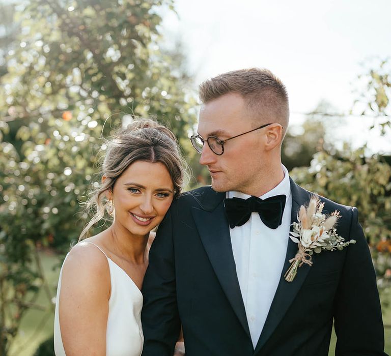 Bride in satin wedding dress leans against the groom in black tie with a dried flower buttonhole 