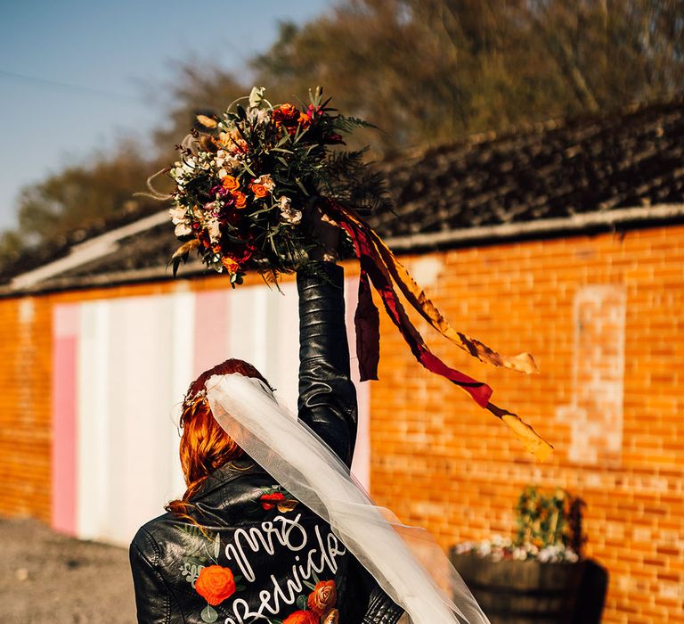 Red and orange wedding flowers and personalised with her new last name wearing a veil and holding her bouquet in the air 