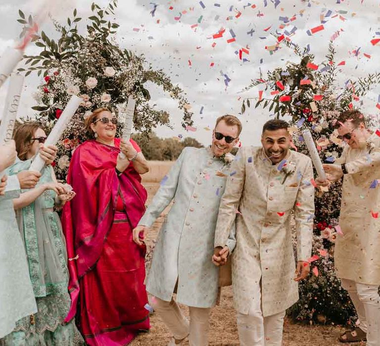 Groom in cream and gold Indian sherwani and white garden rose boutonniere holding hands with groom in mint green Indian Sherwani with white garden rose and foliage boutonniere and grooms sunglasses doing colourful wedding confetti canon exit 