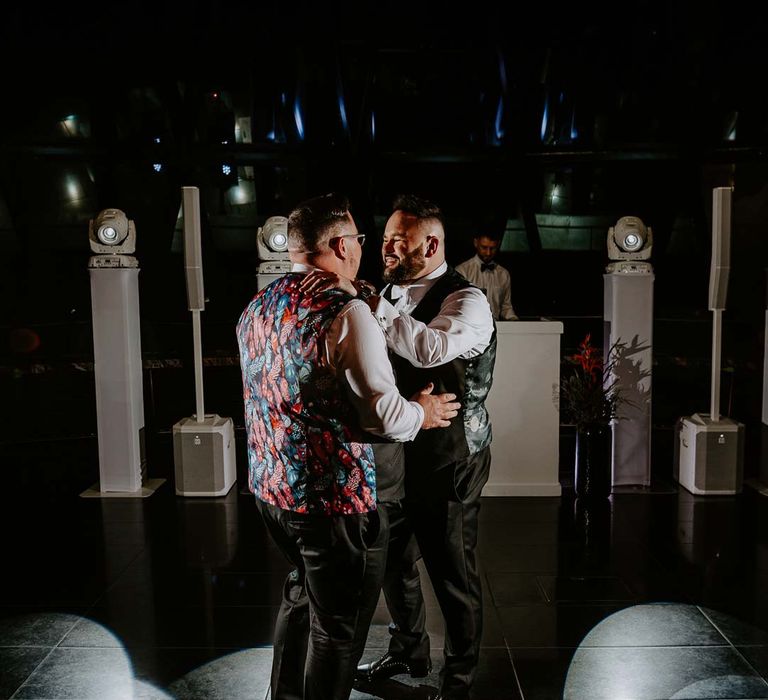 Grooms in matching black waistcoats with colourful patterned backs, white shirts and black suit trousers dancing at their nightclub-style wedding party 