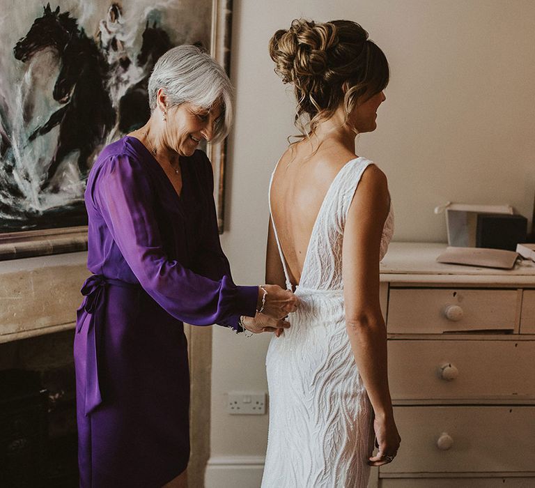 Mother of the bride in vibrant purple dress helps the bride into her form fitting wedding dress with pretty intricate details 